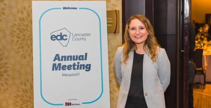 Brianna Titi wears a black shirt and gray blazer as she stands next to a sign that says, "EDC Lancaster County Annual Meeting Breakfast."